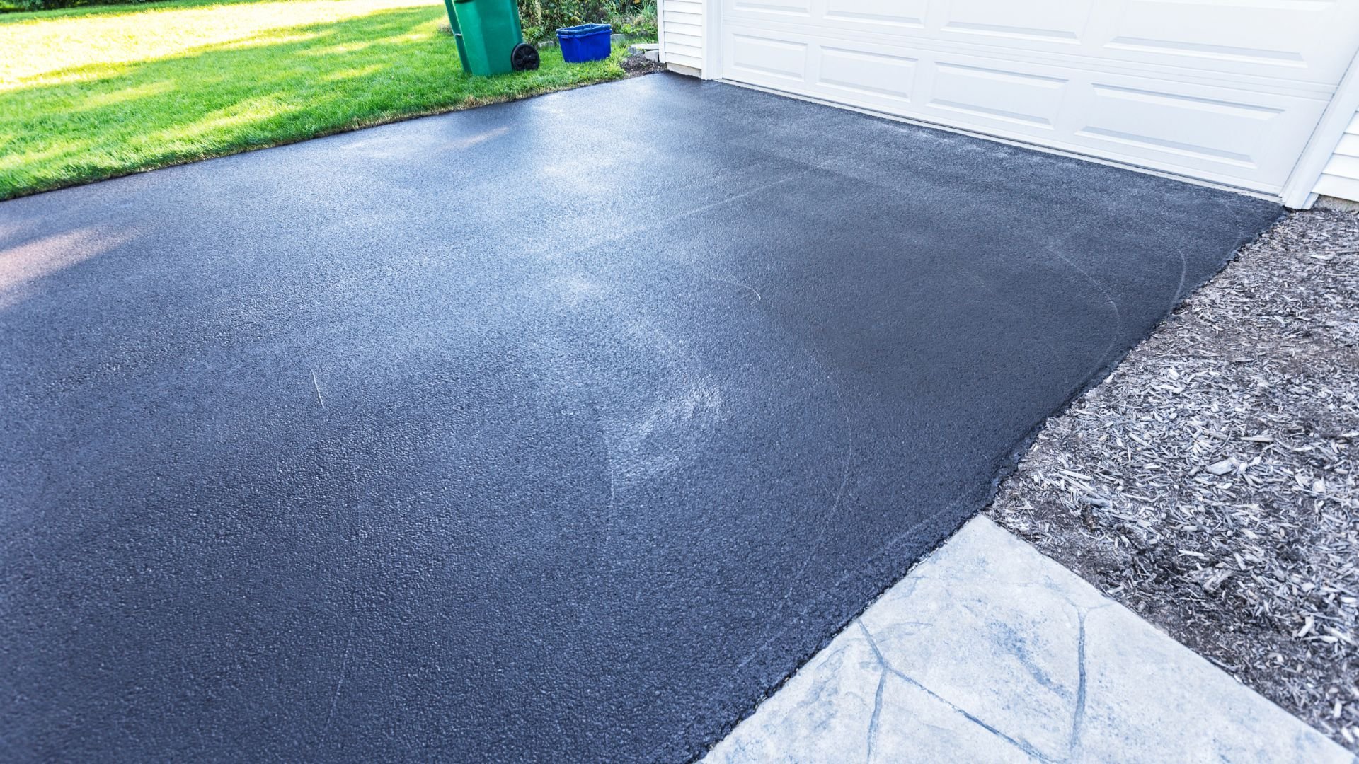 Newly sealed gray asphalt driveway next to green lawn and white garage